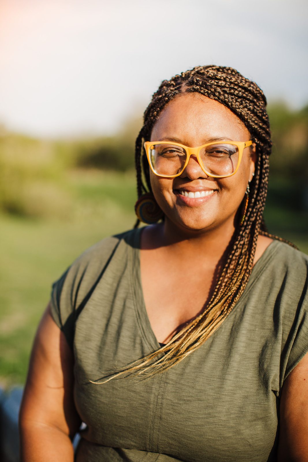 Acelli Crippen-Kok smiling, in yellow glasses and an olive-green top, braids over one shoulder, with a soft green field behind her.