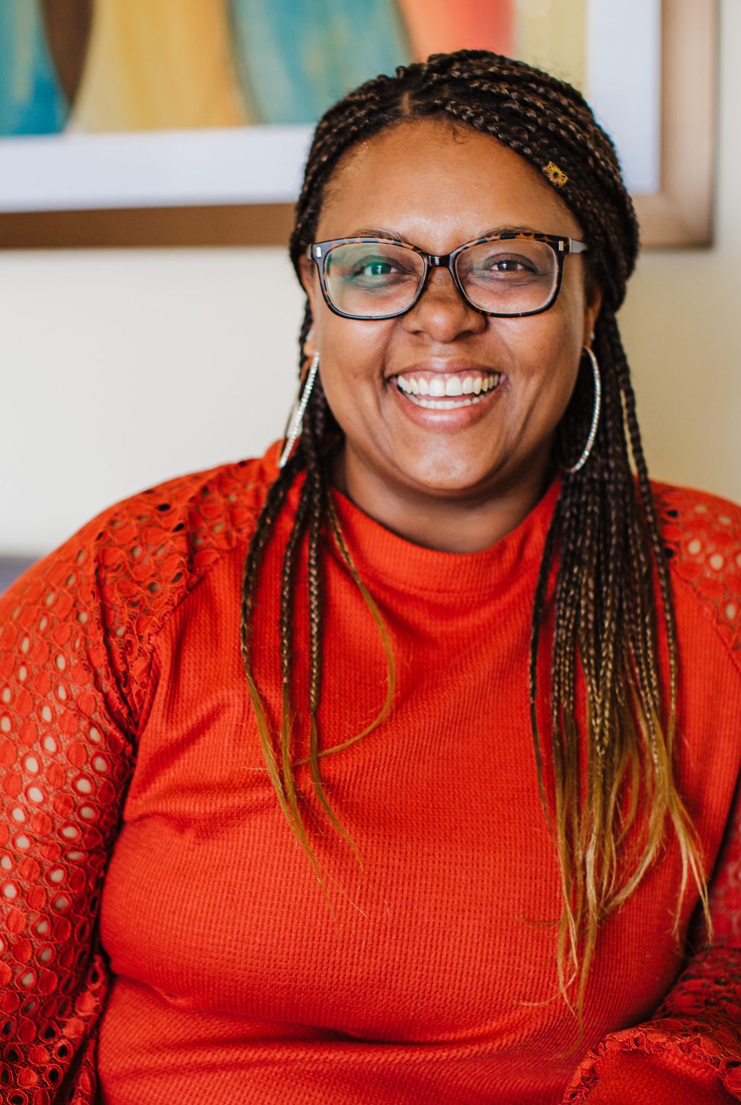 Acelli Crippen-Kok laughing, in tortoiseshell glasses and a rust-orange top with lace sleeves, braids down past her shoulders, a bright painting behind her.