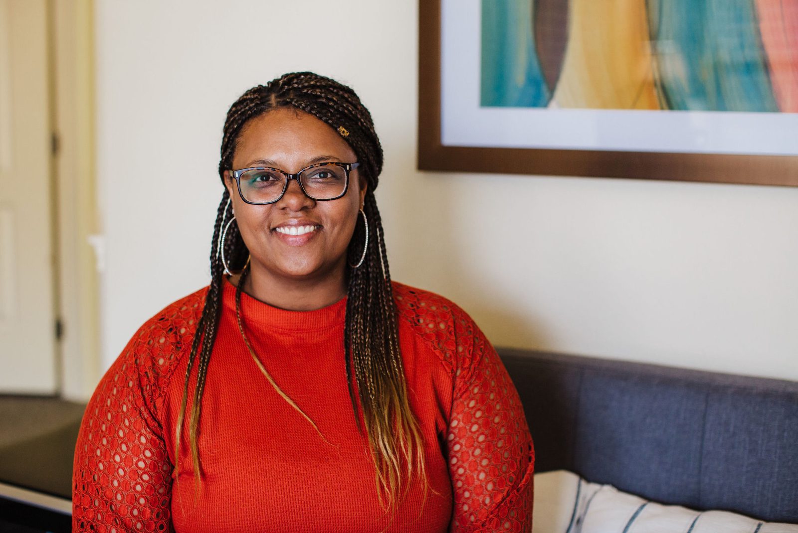 Acelli Crippen-Kok standing in her office, smiling, in tortoiseshell glasses and a rust-orange top with lace sleeves, her braids down past both shoulders, a cream wall behind her.