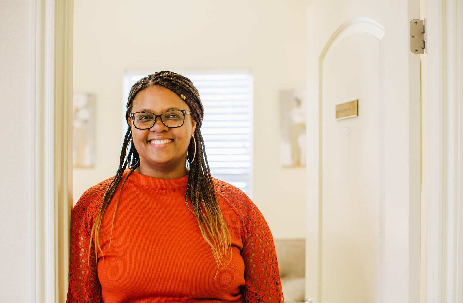 Acelli Crippen-Kok standing in the open doorway of her office, smiling, in tortoiseshell glasses and a rust-orange top with lace sleeves, her braids down past her shoulders, a bright room with window blinds behind her.