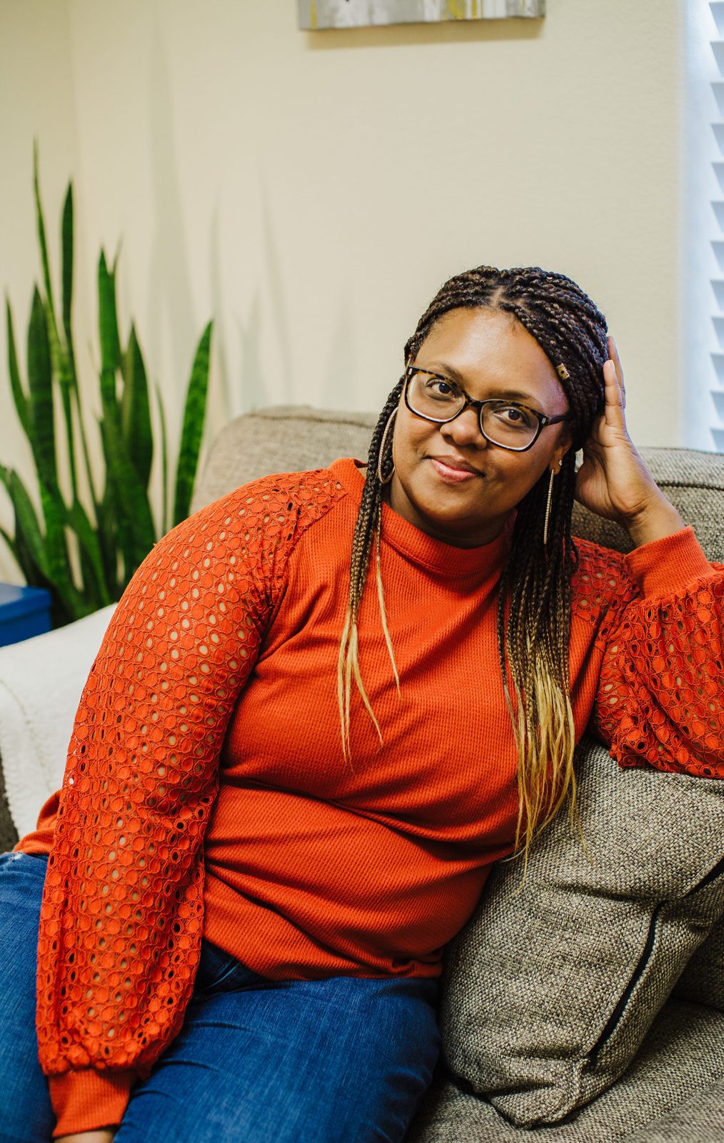 Acelli Crippen-Kok seated on a grey couch in her office, in tortoiseshell glasses and a rust-orange top with lace sleeves, one hand to her head, a snake plant behind her.