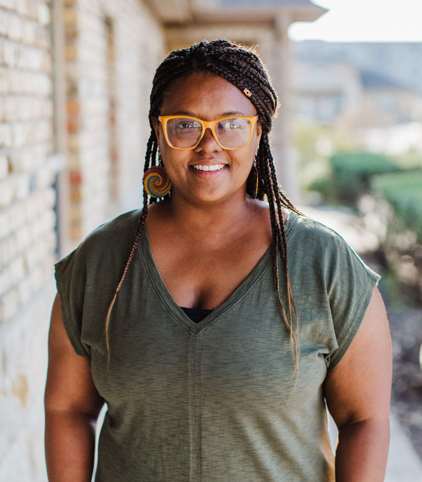 Acelli Crippen-Kok standing on a walkway outside, smiling, in orange-framed glasses, long braids and an olive green top, a pale stone wall behind her.