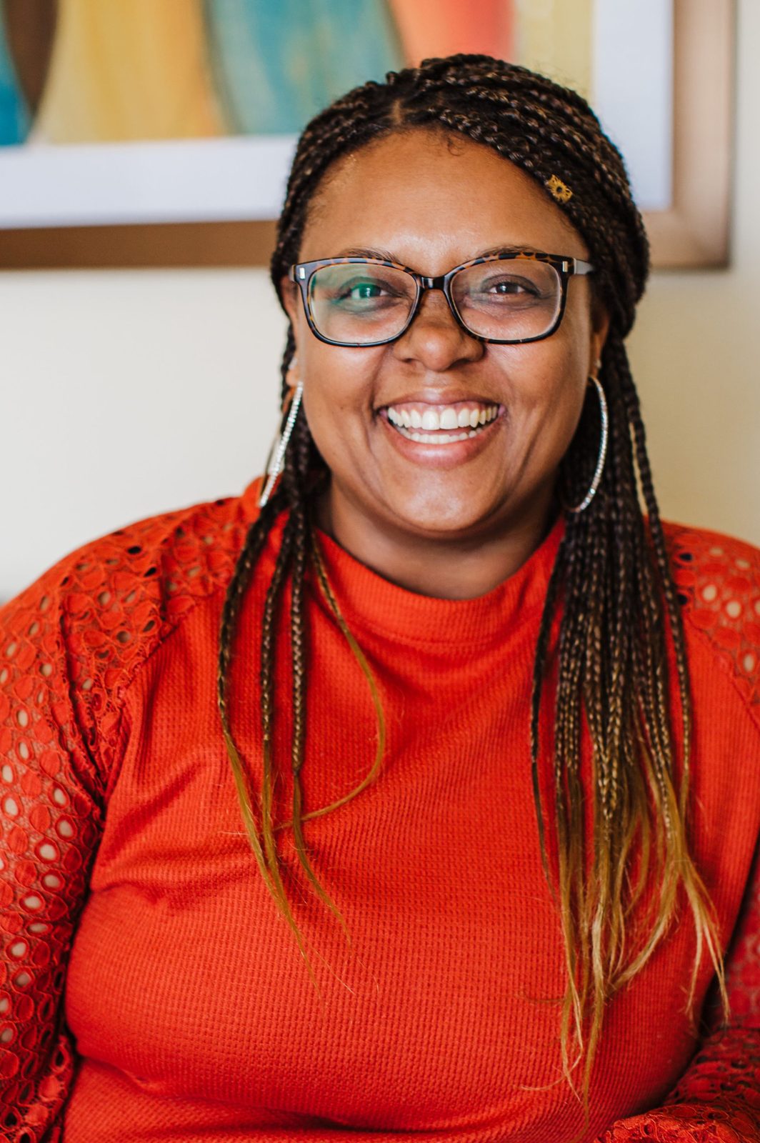 Acelli Crippen-Kok smiling in her office, in tortoiseshell glasses, hoop earrings and a rust-orange top with lace sleeves, long braids over both shoulders, a bright painting on the wall behind her.